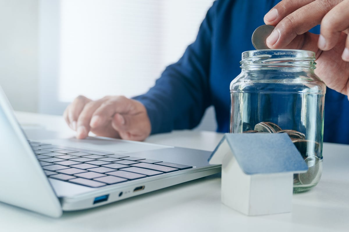 Holding a house model beside a savings jar and laptop, symbolizing planning for a home sale